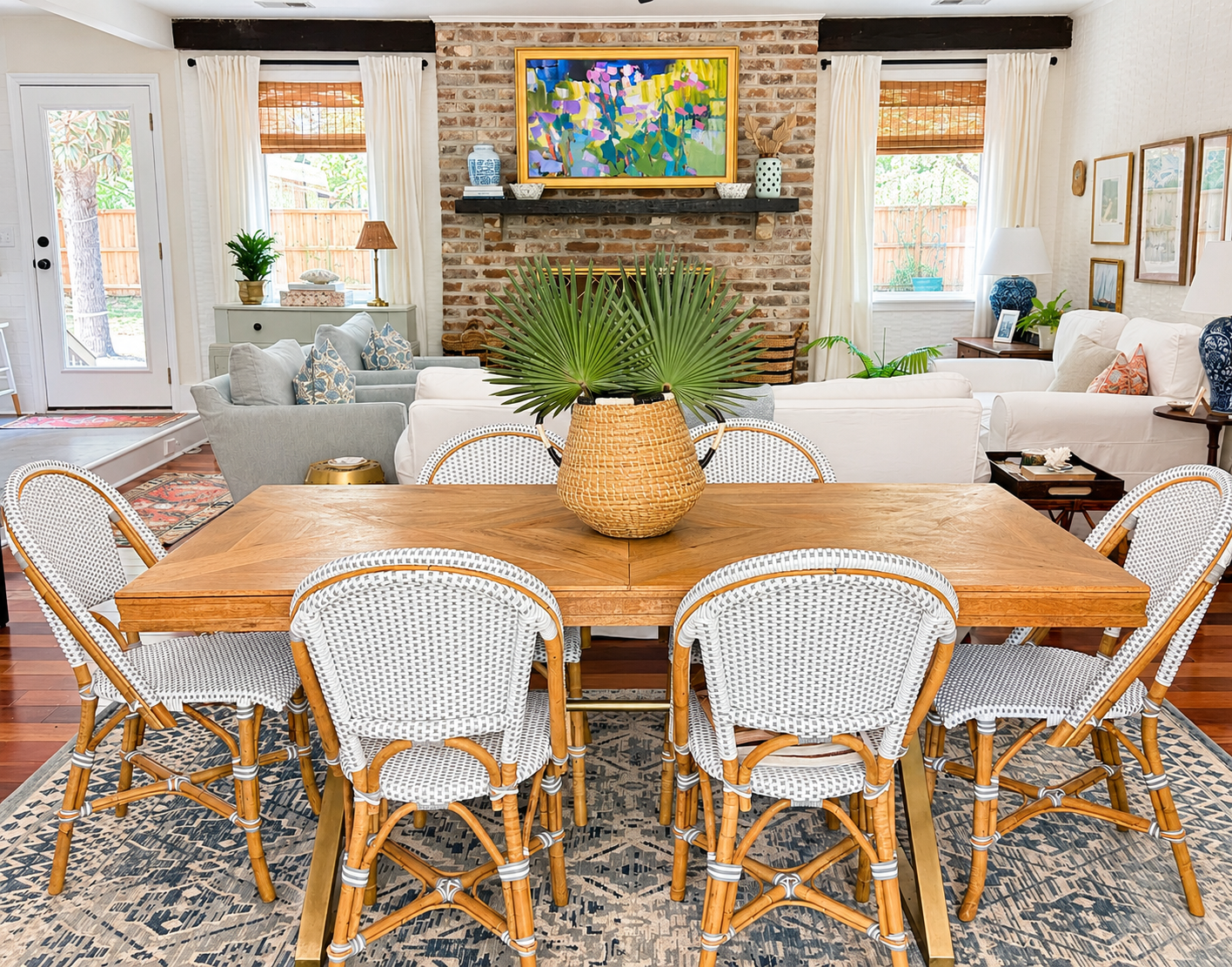 Dining table and woven chairs with a brick fireplace and colorful floral artwork beyond