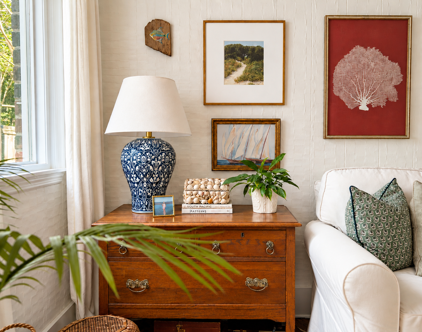 Antique wood chest with a blue-and-white lamp, layered artwork, plant, and white chair with a green patterned pillow