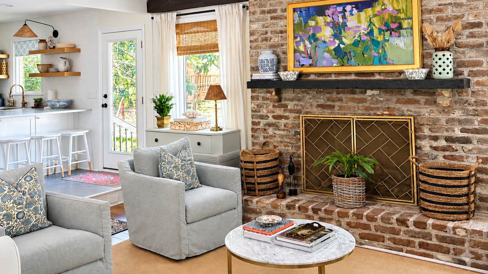 Collected living room with a brick fireplace, colorful floral artwork, brass fireplace screen, blue-gray chairs, and a round marble coffee table, opening onto the kitchen
