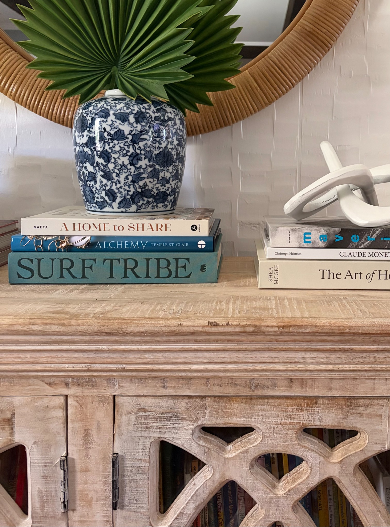 Blue-and-white porcelain vase with palm leaves, a stack of books, and a pale wood cabinet beneath a rattan mirror
