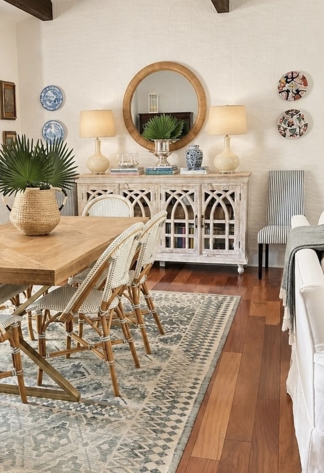 Angled dining room with a round rattan mirror, two lamps, pale wood cabinet, woven dining chairs, and a patterned rug